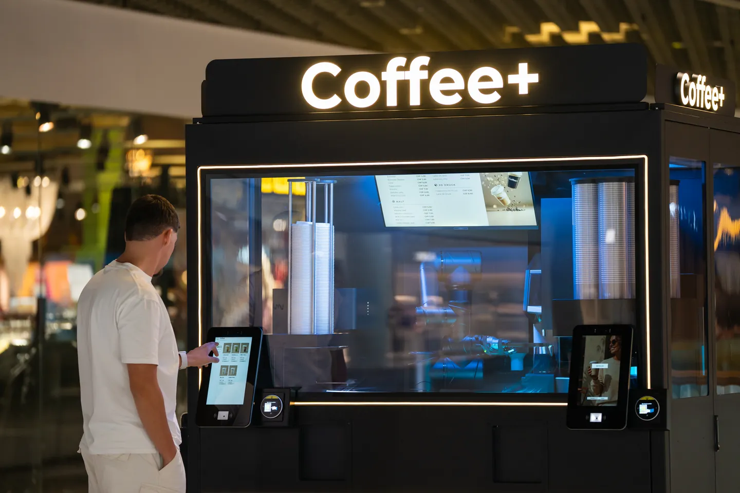 Ein Gast wählt sein Getränk am linken Display des Coffee+ Kiosks. Die beleuchtete Anlage und der Roboterarm sind hinter Glas sichtbar.
