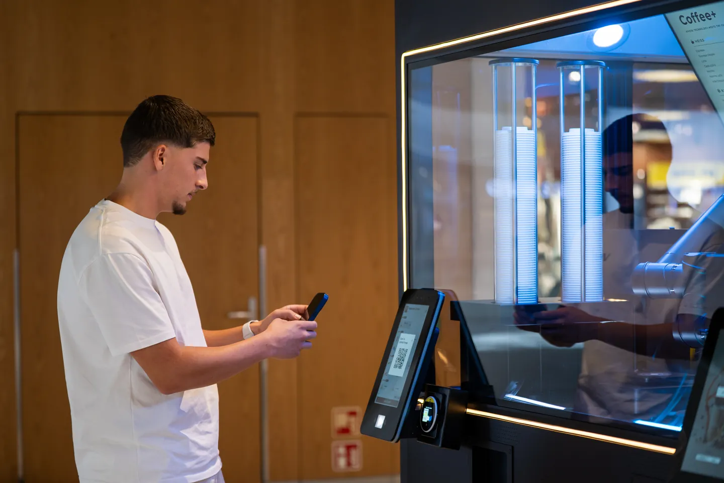 Ein Gast hält sein Smartphone vor das Bestelldisplay des Coffee+ Kiosks, auf dem ein QR-Code angezeigt wird.