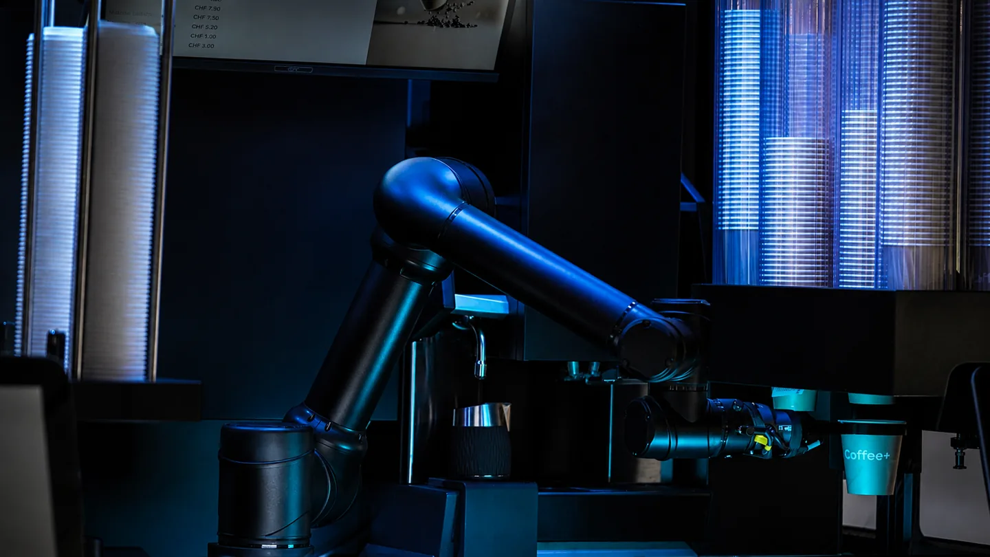 Der Roboterarm bei der Zubereitung, zwischen Becherstapeln, Milchkanne und einem Coffee+ Becher.